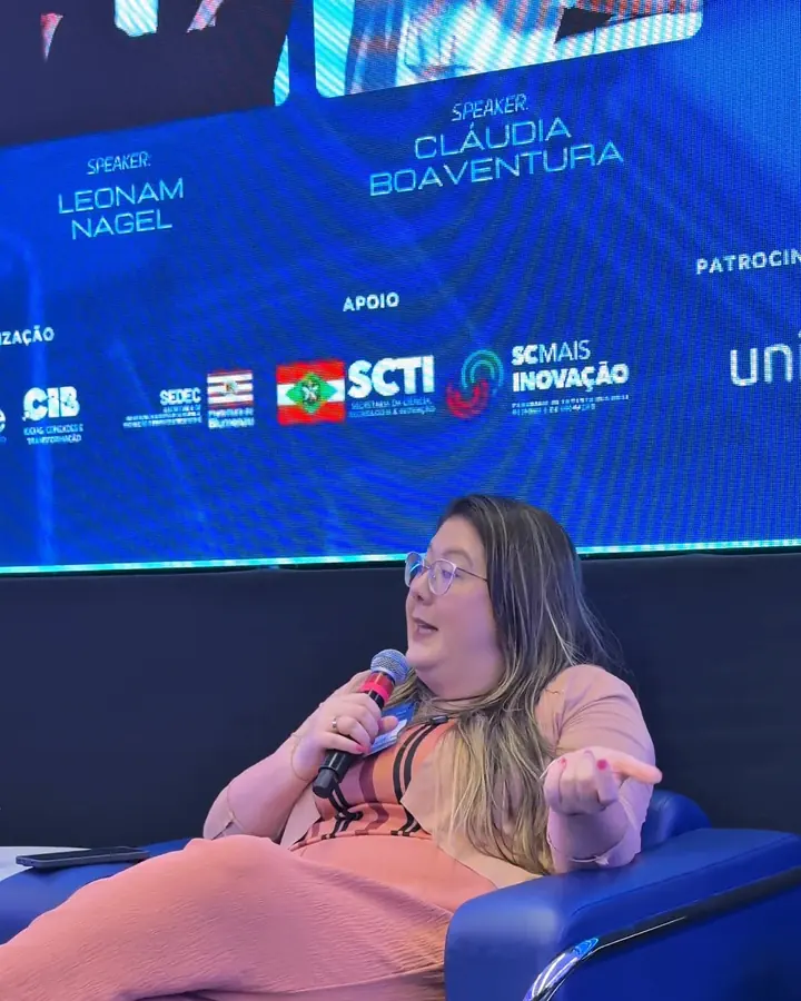 Clau Boaventura com o microfone num painel sobre IA da SCTI, sentada numa poltrona azul