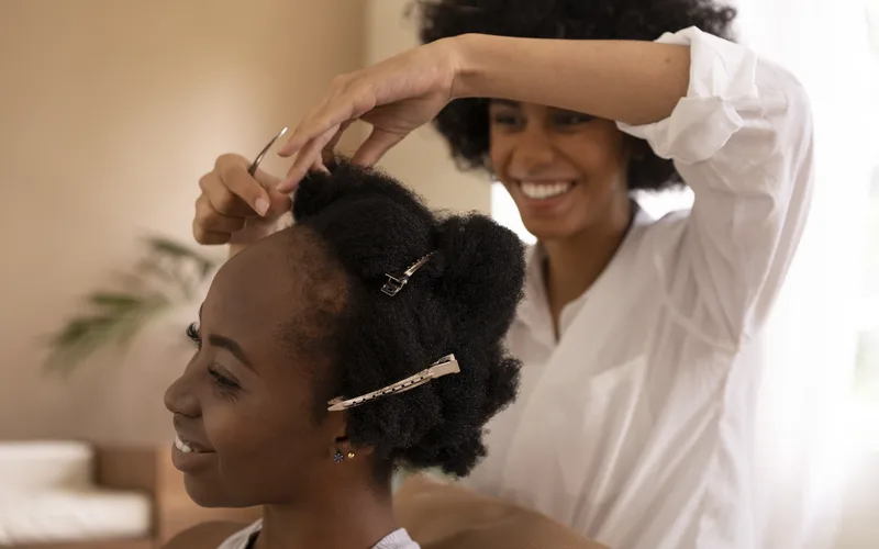 Cabeleireira finalizando o penteado de uma cliente loira e sorridente num salão claro e clean.