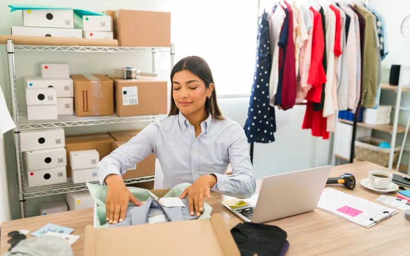 Dona de loja virtual de blusa vermelha conferindo pedidos na prancheta, entre caixas de papelão e uma arara de roupas coloridas.
