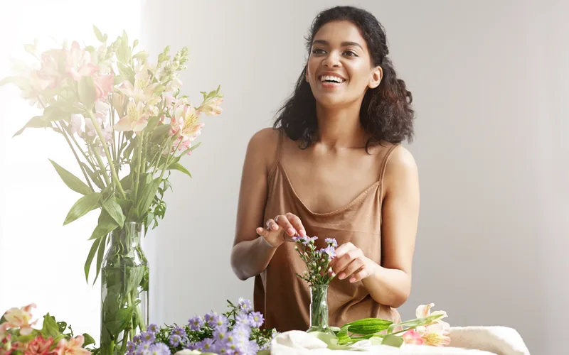Florista sorridente de avental xadrez montando um arranjo de flores coloridas num ateliê claro.