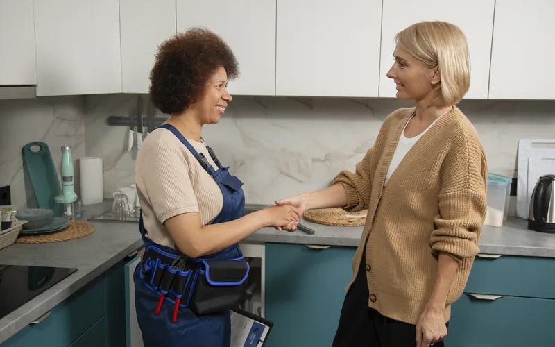 Encanadora de macacão cumprimentando a cliente numa cozinha clara, depois do conserto.