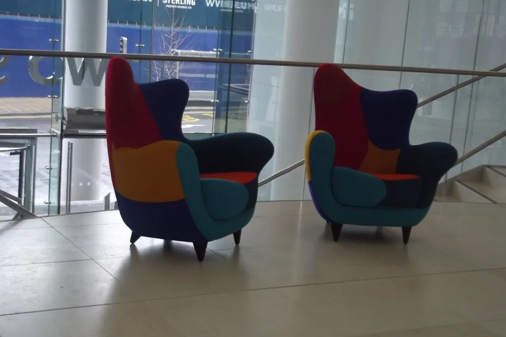 Duas poltronas coloridas, uma de frente para a outra, num saguão claro com parede de vidro.