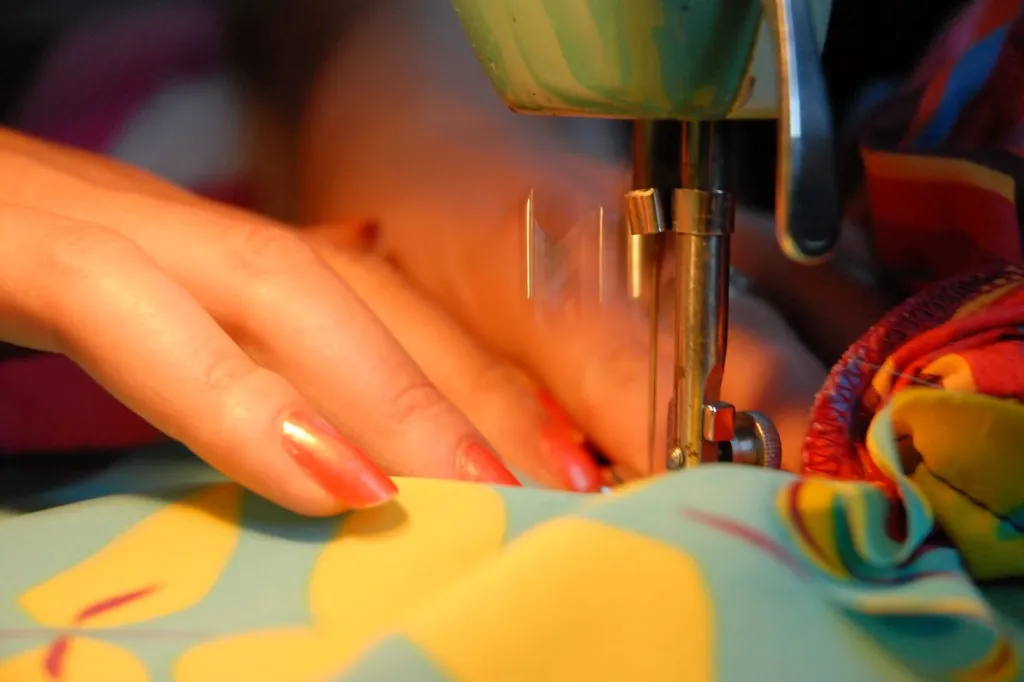 Mãos de uma costureira guiando um tecido estampado sob a agulha da máquina de costura.