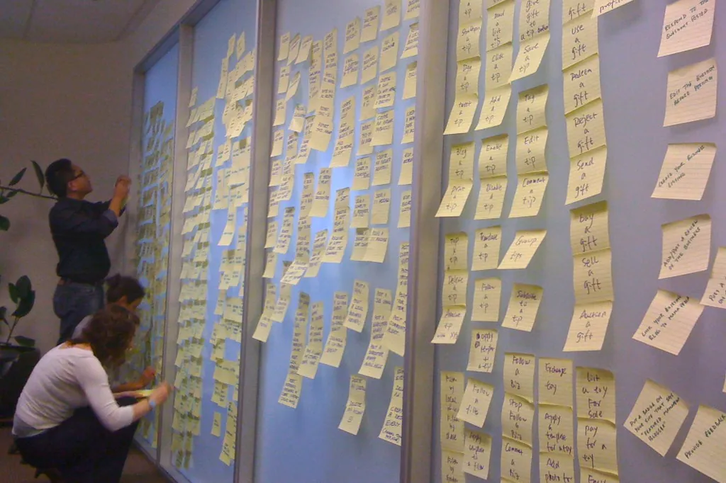 Duas pessoas organizam centenas de post-its numa parede de vidro durante uma sessão de planejamento.
