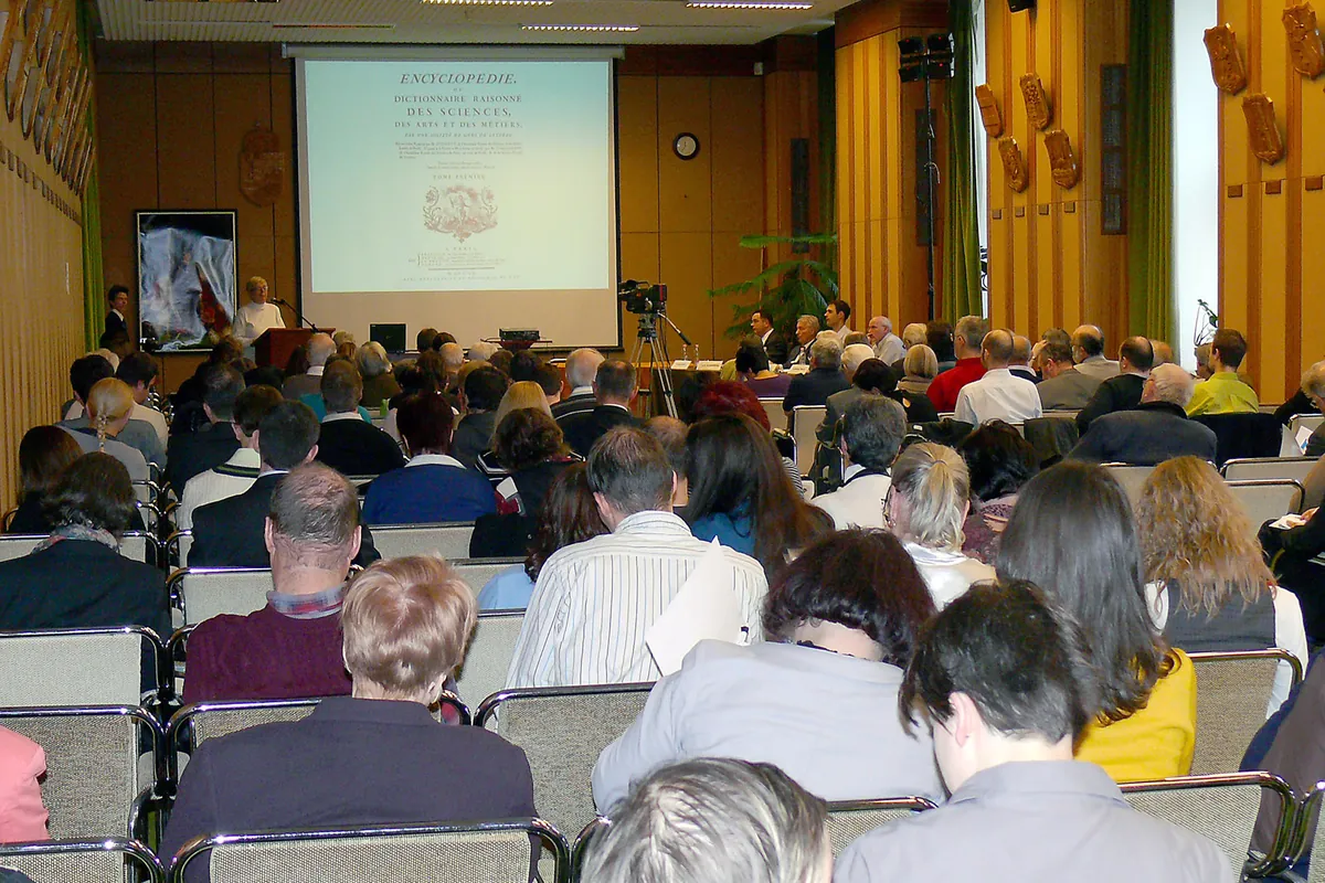 Plateia vista de trás num auditório de madeira, atenta à apresentação projetada no telão à frente.