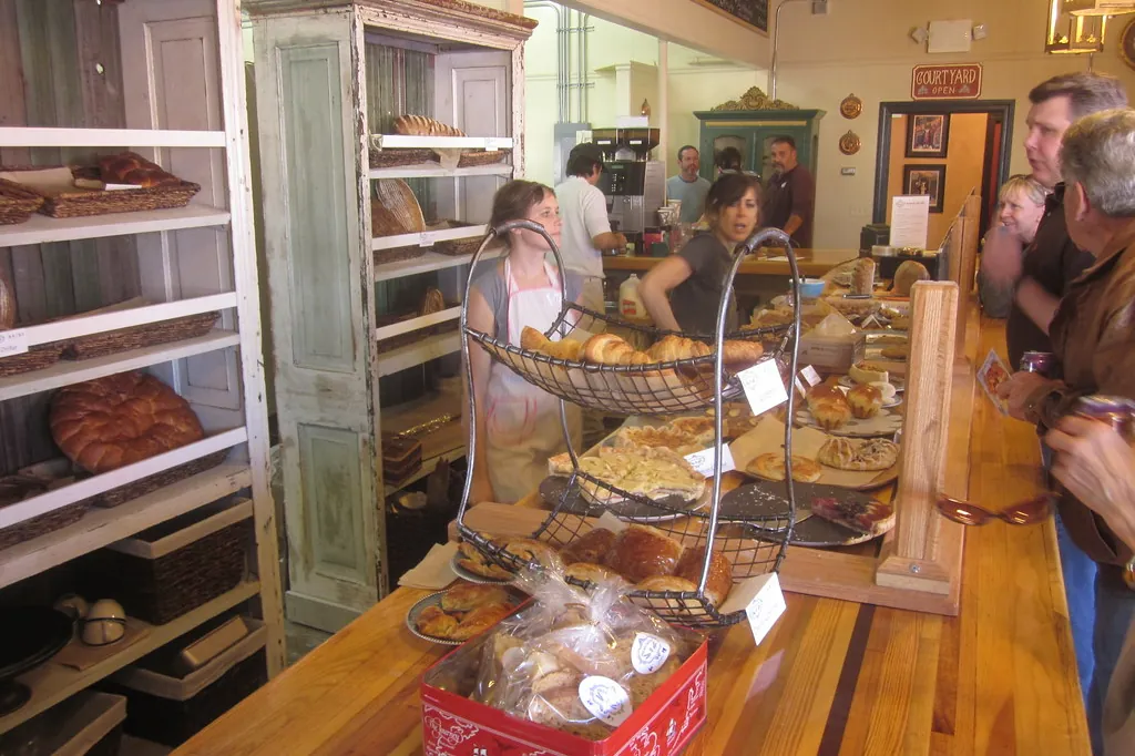 Balcão de madeira de uma padaria de bairro, com cestas de pão e clientes sendo atendidos ao fundo.