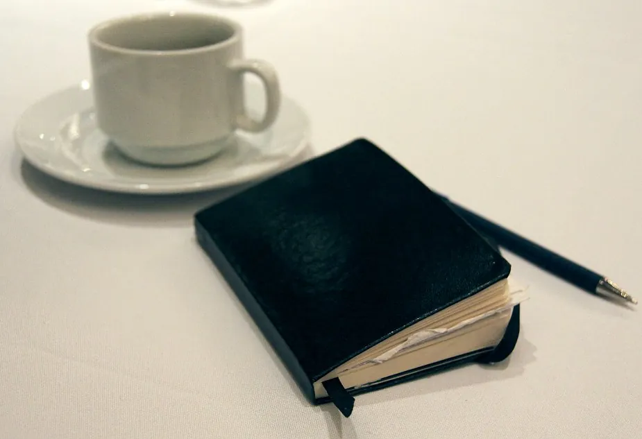 Xícara de café, caderno de capa preta e caneta sobre uma mesa clara.